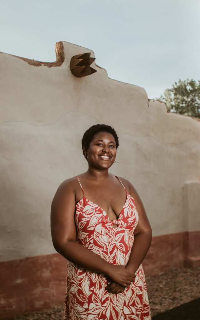 Kiana Elkins 2022 A photo of a Black woman posing for a photo in front of a tan clay wall, her hands are crossed at the waist and she is smiling